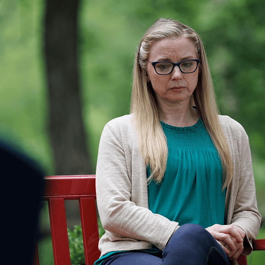 A white woman with long blonde hair and dark glasses looking down, frowning. She is sitting and seen from the waist up