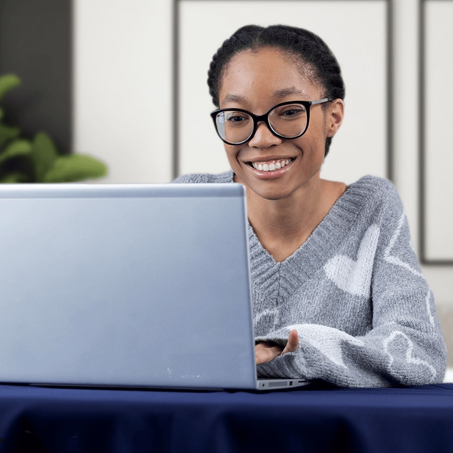 A young African-American woman with braided hair and dark glasses smiles while typing on a laptop