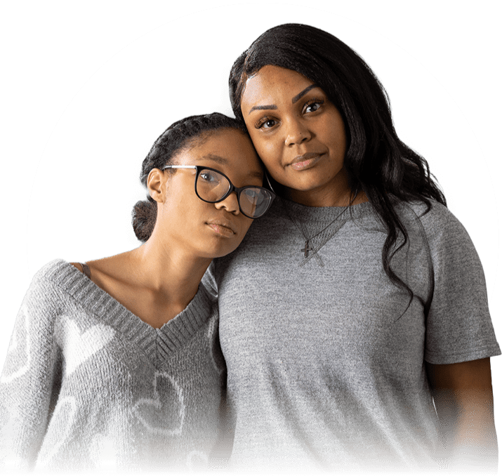 A young African-American girl in dark glasses leans on an older African-American woman with long brown hair. The pair are seen from the waist up and they both have neutral expressions
