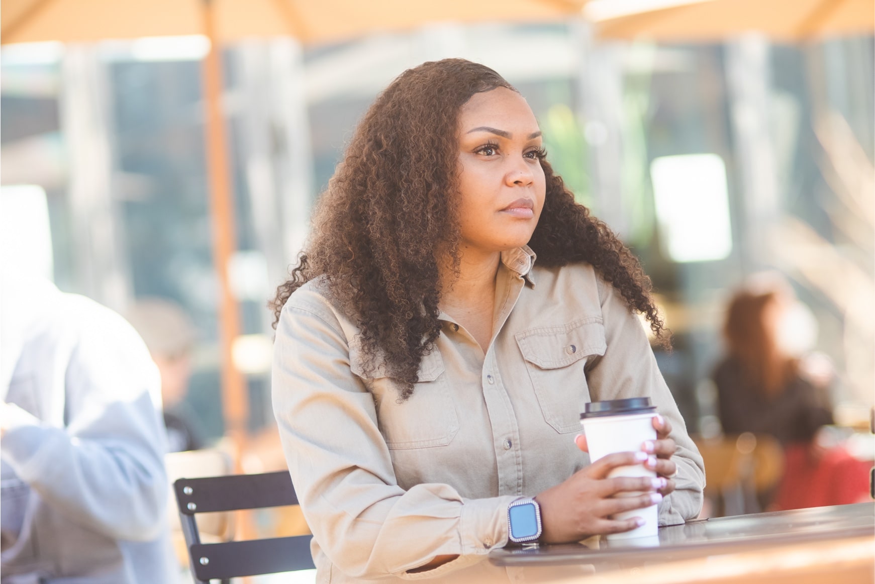 An African-American woman with long brown hair sits outside at a table. She is holding a white coffee cup, and she is looking far off in the distance
