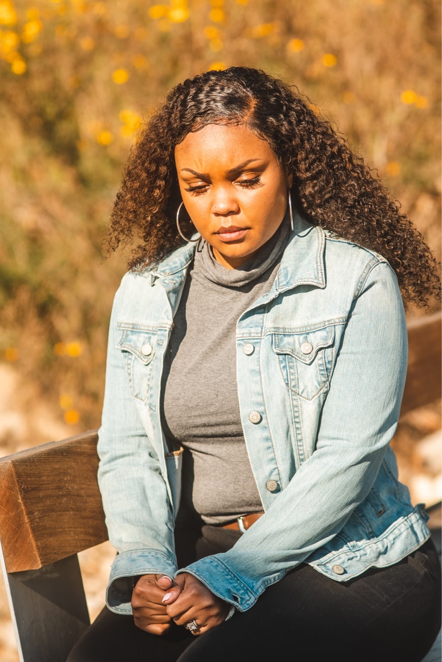 An African-American woman with long brown hair sits outside on a wooden chair. Her hands are folded in front of her, and she is looking down, away from the camera