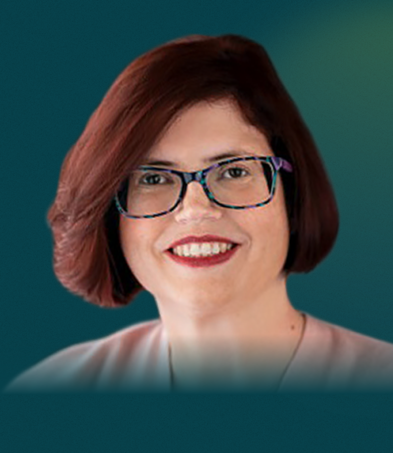 A white woman with short brown hair and patterned glasses is seen from the neck up. She faces the camera with a smile that reveals her teeth