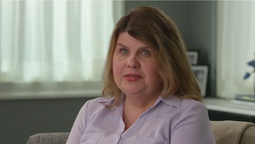 Video still of a white woman with shoulder-length dirty blonde hair seen from the neck up. She faces the camera with a neutral expression. The triangular video player button sits on top of the video