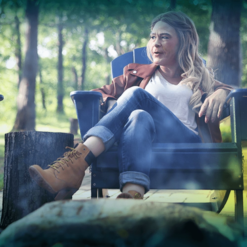 A white woman with long blonde hair is seen sitting in a wooden chair outside. She looking to the right and is in mid-sentence with someone off camera.