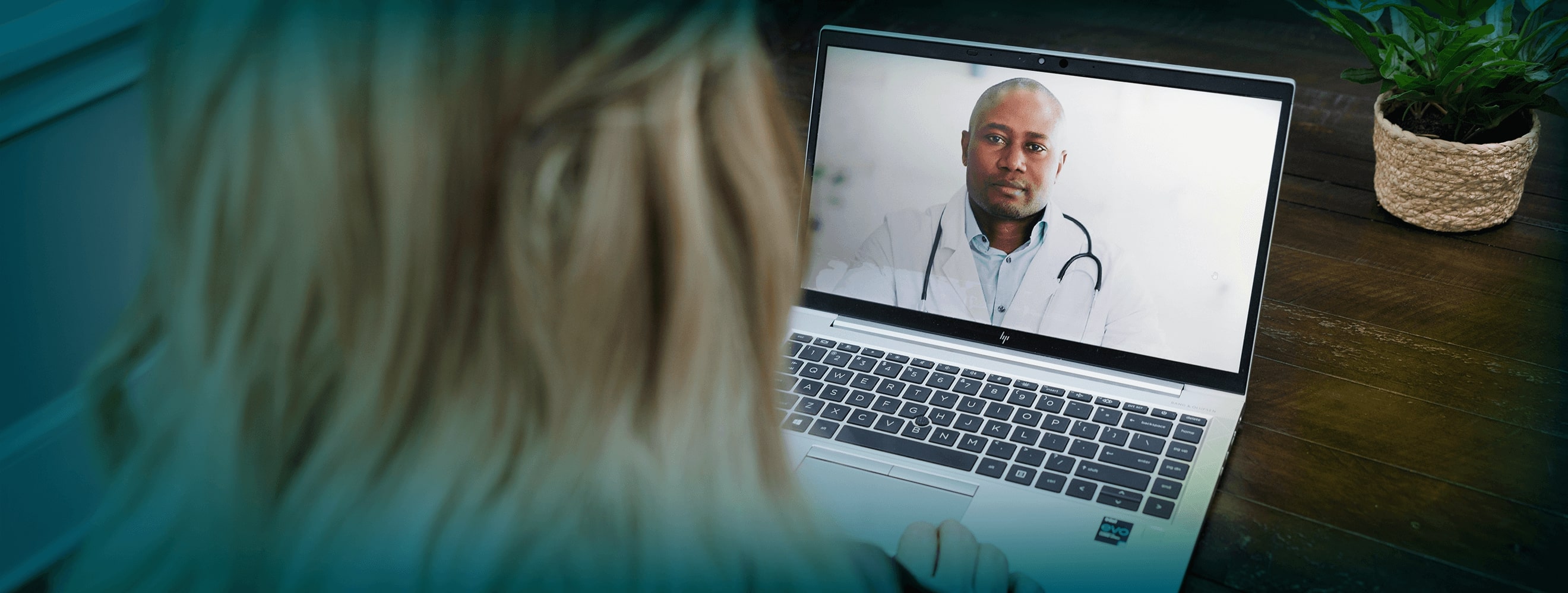 A woman with long blonde hair is seen from the back. An open laptop faces the camera. On the screen is an African- American doctor in a white lab coat with a stethoscope around his neck. He has a neutral expression