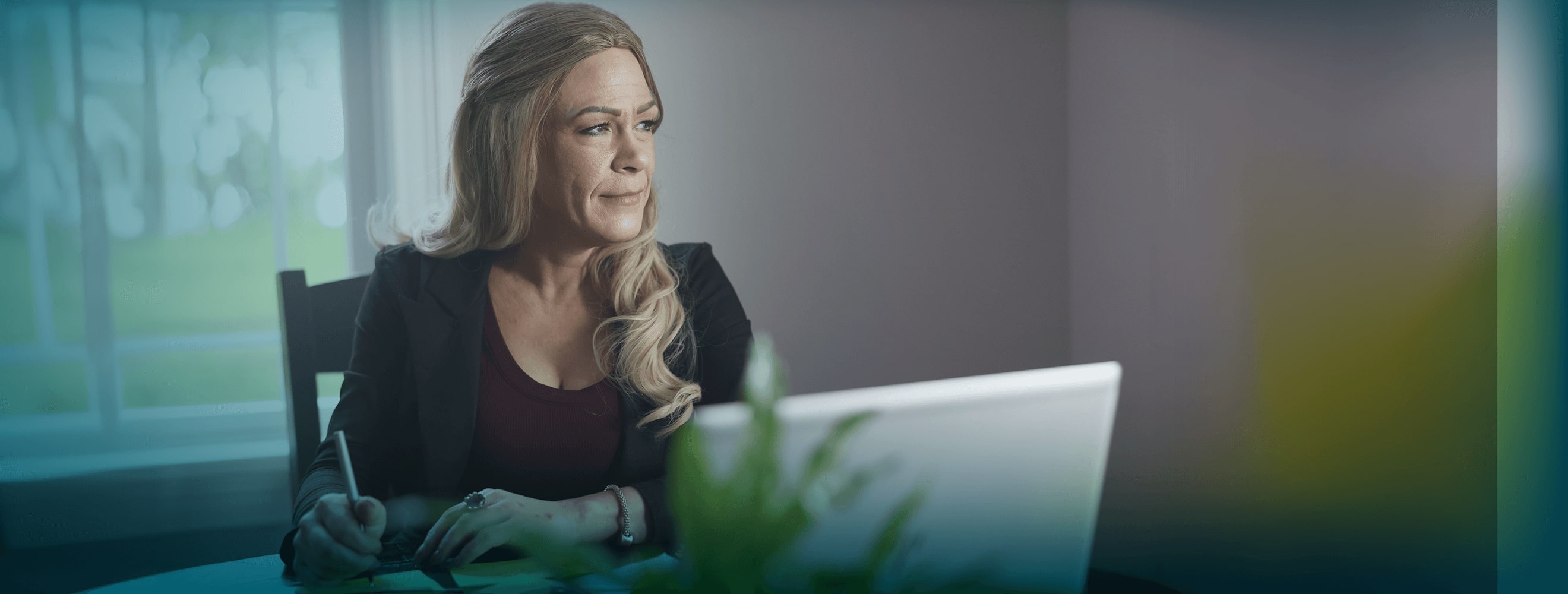 A white woman with long blonde hair is seated at a table. She is holding a pen, in the middle of writing, and is looking off-camera to the right.