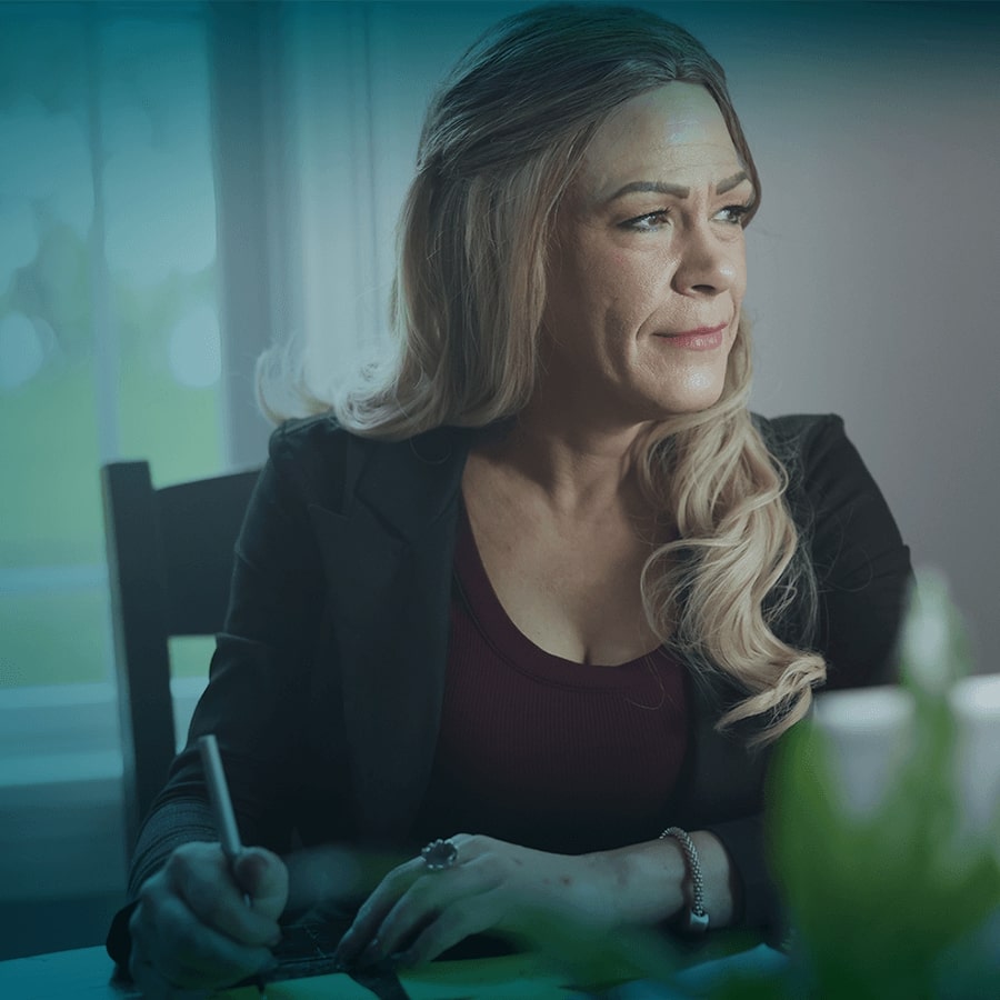 A white woman with long blonde hair is seated at a table. She is holding a pen, in the middle of writing, and is looking off-camera to the right.