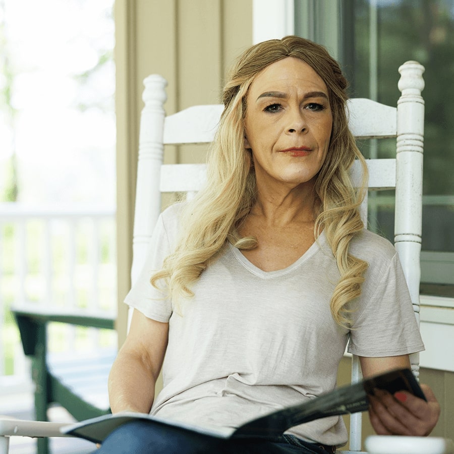 A white woman with long blonde hair. She is seated in a white rocking chair and is looking straight at the camera with a neutral expression.