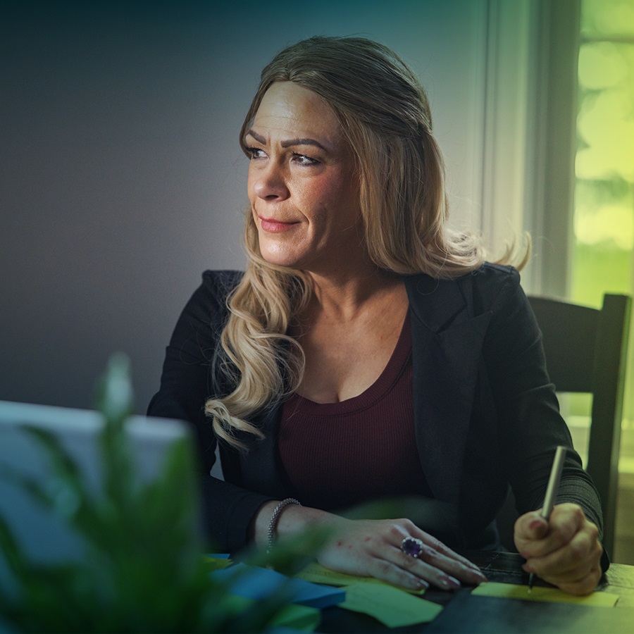 A white woman with long blonde hair is seen from the waist up. She is seated at a table and looking off camera towards the left. In front of her is a laptop, and she’s holding a pen