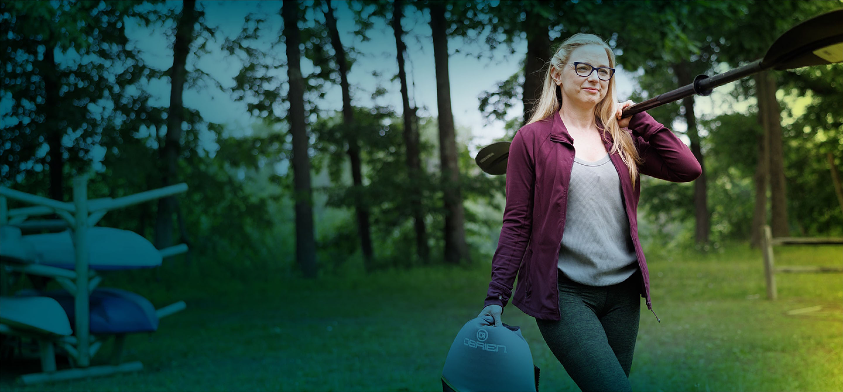 A white woman with black-framed glasses and long blonde hair is seen from the knees up. She is carrying a kayak ore over her right shoulder and holding a wetsuit in her left hand. She has a smile on her face as she walks towards the camera.