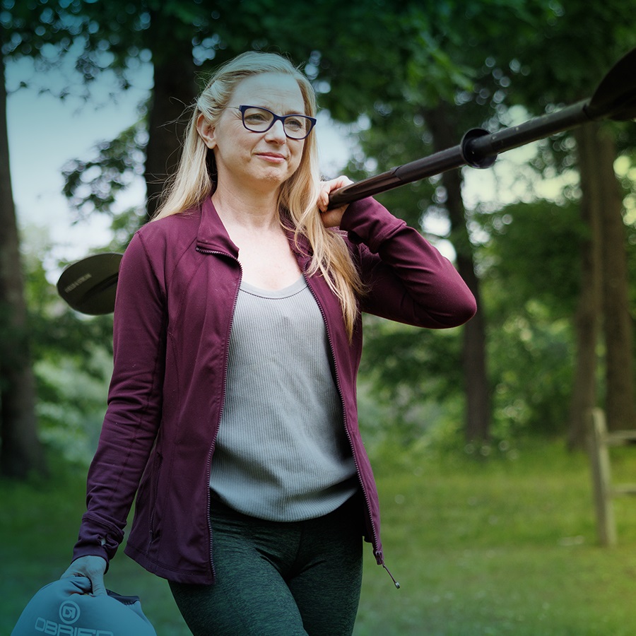 A white woman with black-framed glasses and long blonde hair is seen from the knees up. She is carrying a kayak ore over her right shoulder and holding a wetsuit in her left hand. She has a smile on her face as she walks towards the camera.