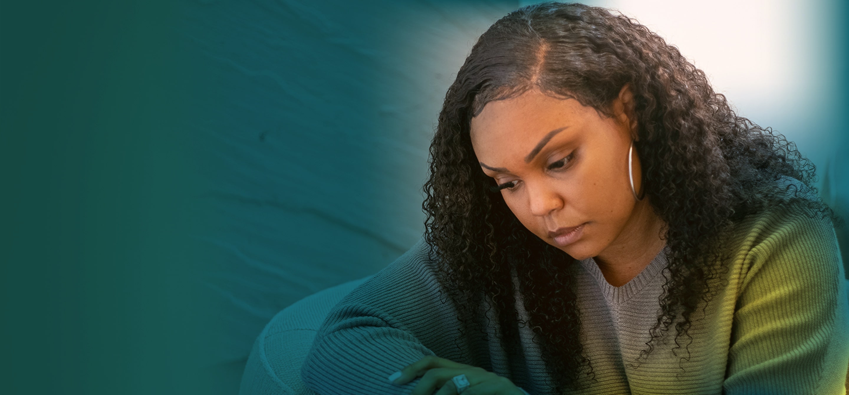 An African-American woman with long brown hair is seen from the chest up. She is hunched over and looking down with a serious expression