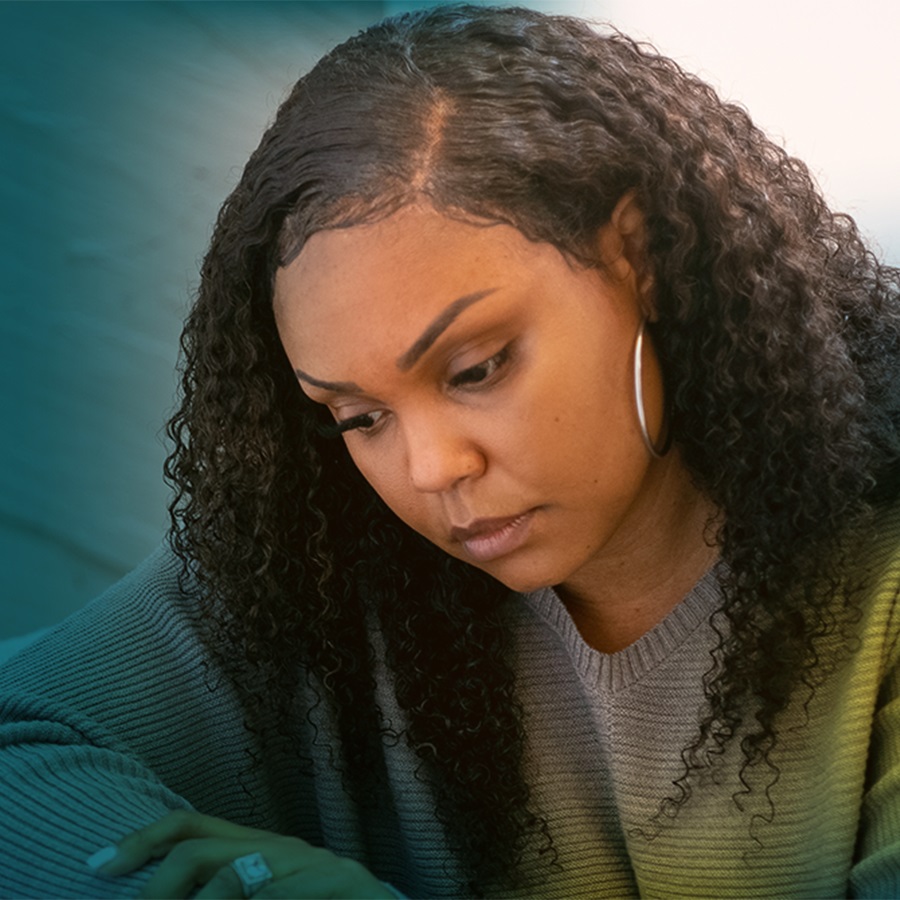 An African-American woman with long brown hair is seen from the chest up. She is hunched over and looking down with a serious expression
