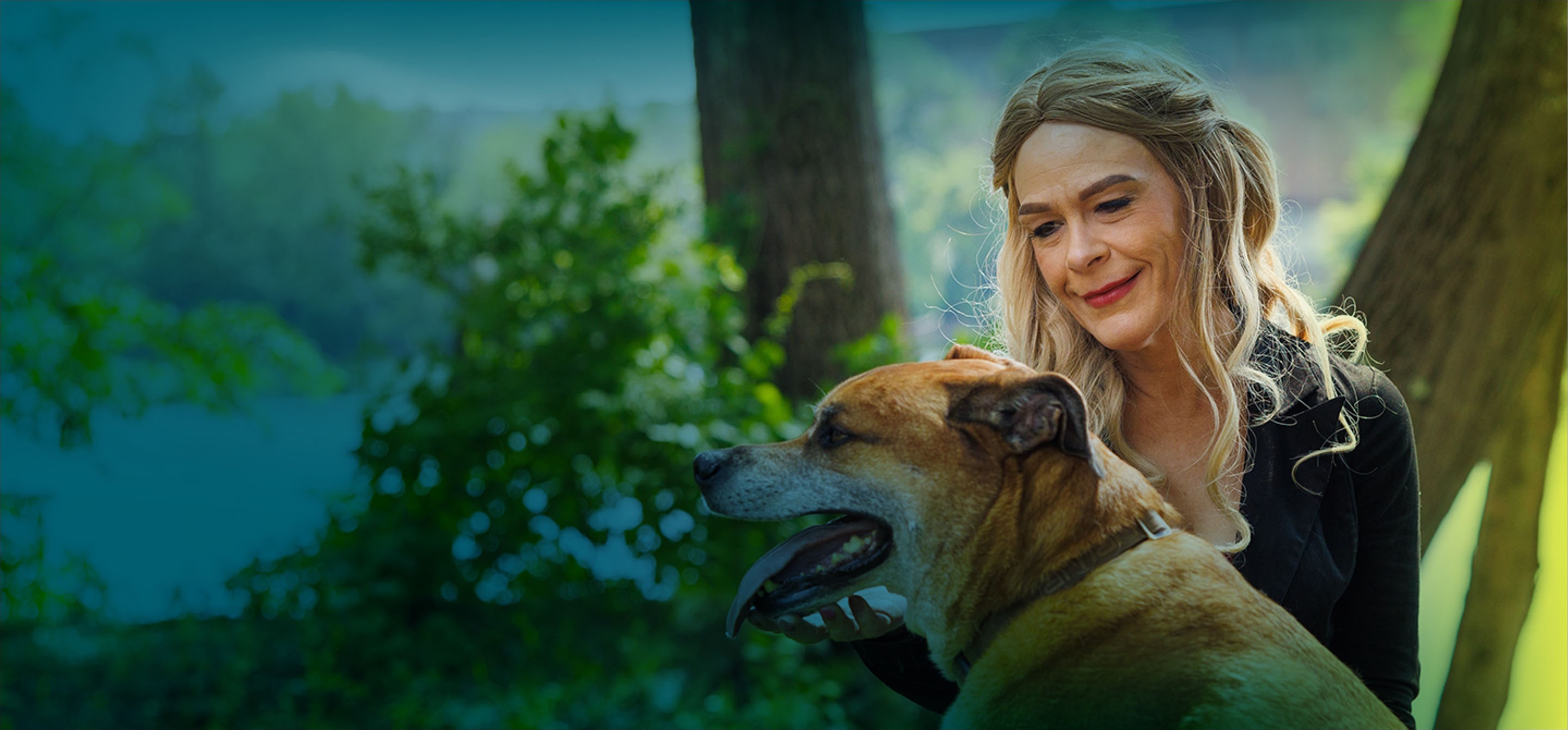 A white woman with long blonde hair is outside, kneeling on the ground, petting a mid-sized dog who has his tongue sticking out.  