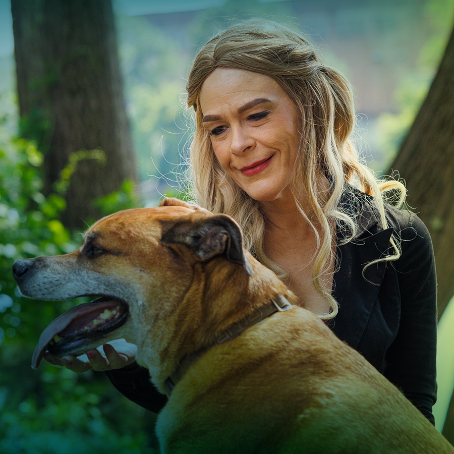 A white woman with long blonde hair is outside, kneeling on the ground, petting a mid-sized dog who has his tongue sticking out.  