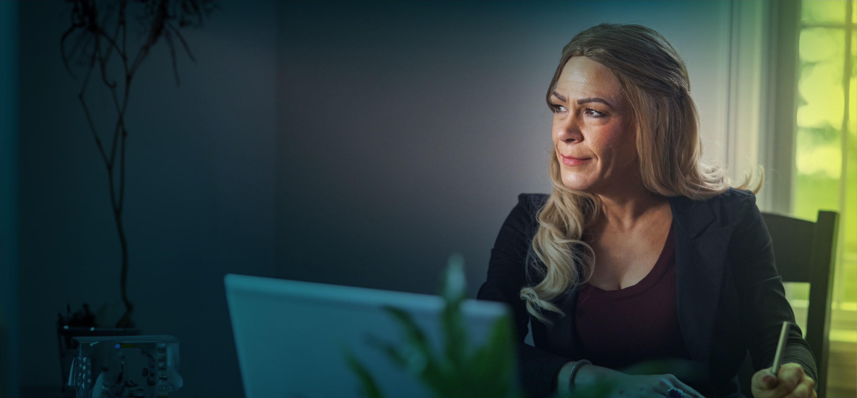 A white woman with long blonde hair is seen from the waist up. She is seated at a table and looking off camera towards the left. In front of her is a laptop, and she’s holding a pen