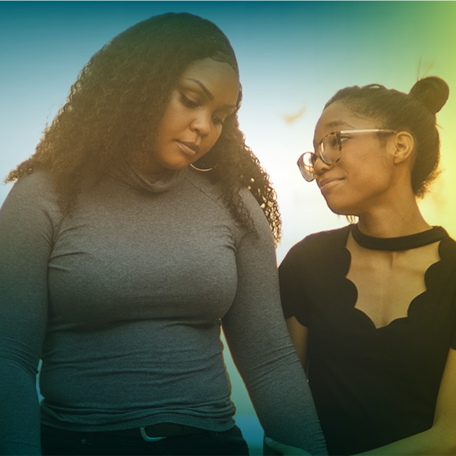 A young African American girl and older African American woman are seen from the waist up. The girl is holding the woman’s hand and looking up at the woman with a look of comfort. The older woman is looking down at the ground with a look of sadness. 