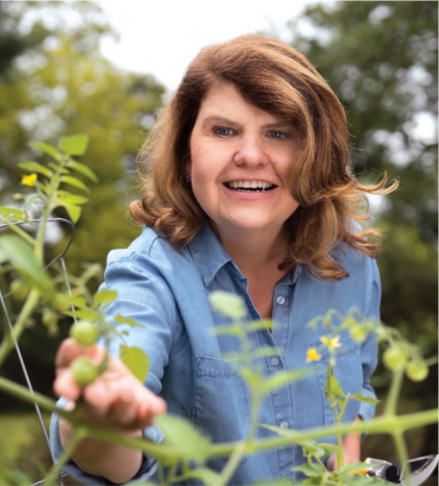 A white woman with shoulder-length dirty blonde hair is seen from the waist up outside. She faces the camera, reaching out her hand to touch a plant. She is smiling, revealing her teeth