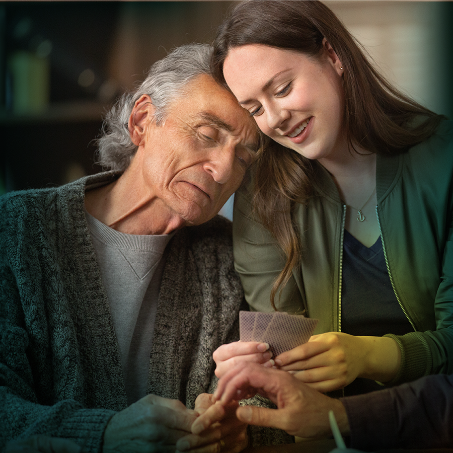 An older white man with gray hair is seen from the waist up leaning against a young white woman with brown hair. The woman is smiling and holding up a hand of cards. The older man has his eyes mostly closed and his mouth turned down.