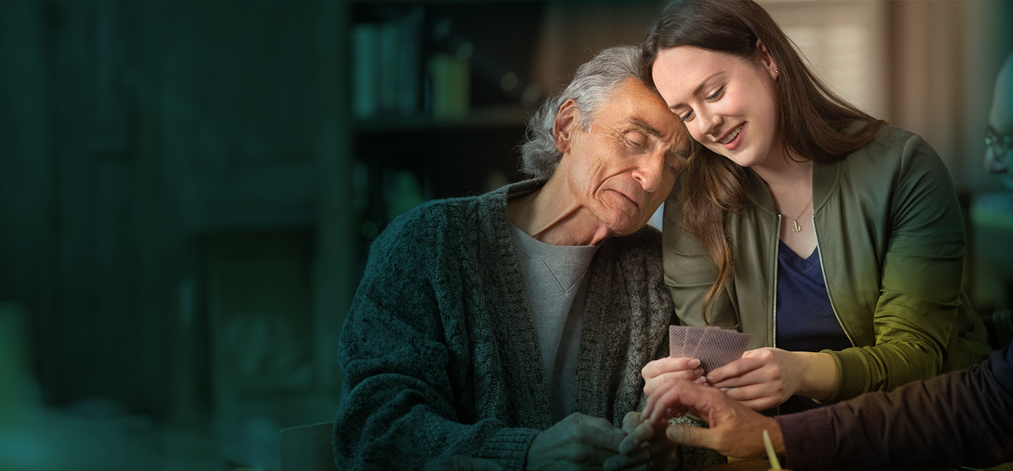 An older white man with gray hair is seen from the waist up leaning against a young white woman with brown hair. The woman is smiling and holding up a hand of cards. The older man has his eyes mostly closed and his mouth turned down.