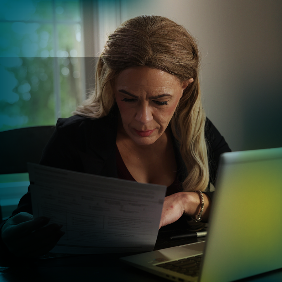 A white woman with long blonde hair is seen from the waist up. She is seated at a table and looking at paperwork. In front of her is an open laptop