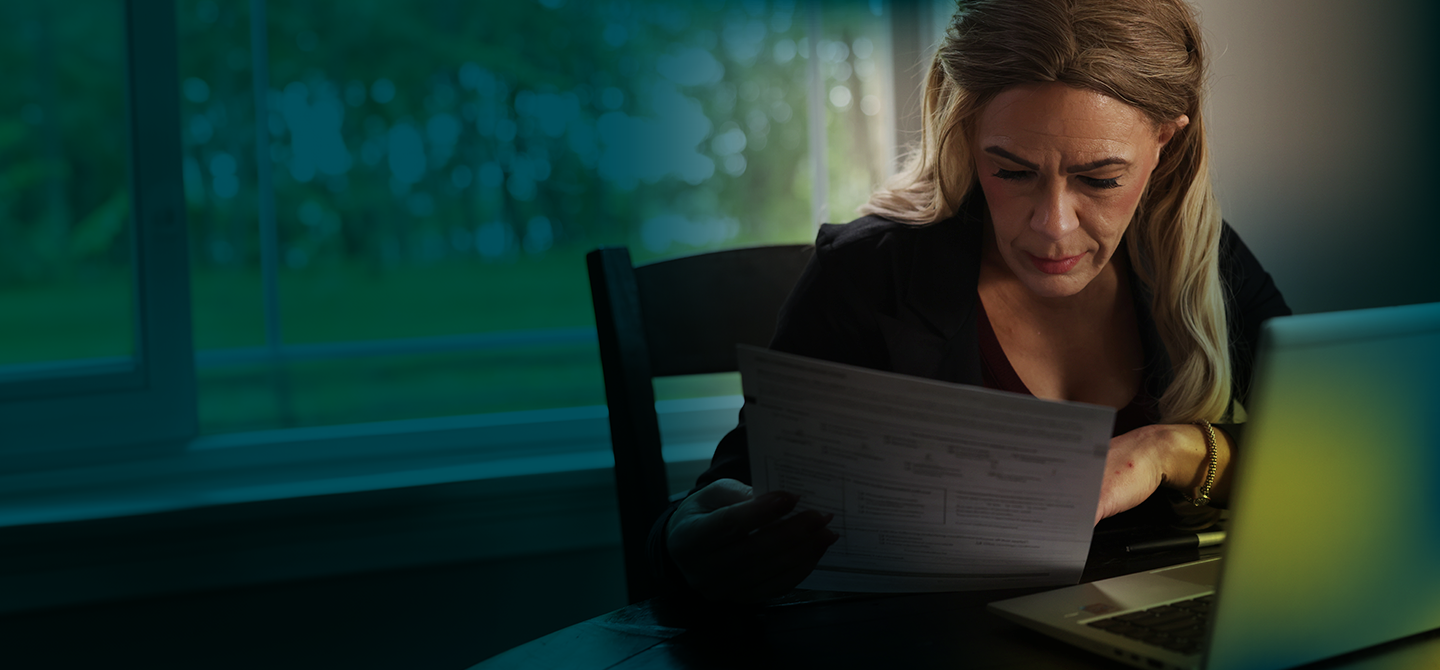 A white woman with long blonde hair is seen from the waist up. She is seated at a table and looking at paperwork. In front of her is an open laptop