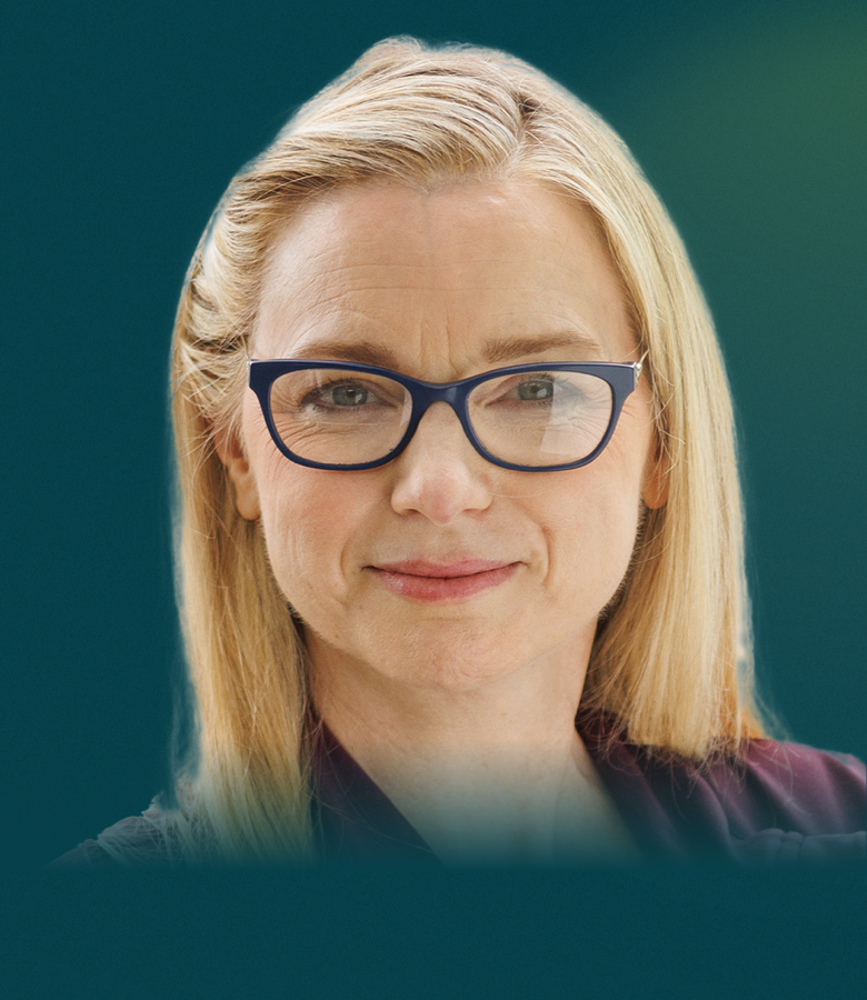 A white woman with long blonde hair and dark glasses is seen from the neck up. She faces the camera with a neutral smile