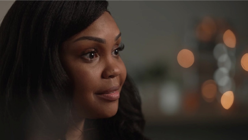 Video still of an African- American woman with long brown hair seen from the neck up. She is seen in profile, looking to the right. The triangular video player button sits on top of the video