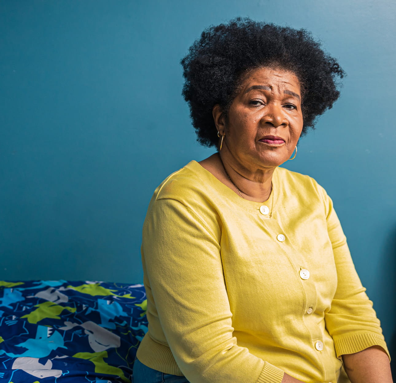 An African-American woman with short, curly black hair is seen from the chest up. She is wearing a yellow cardigan and sitting facing the camera with a neutral expression.