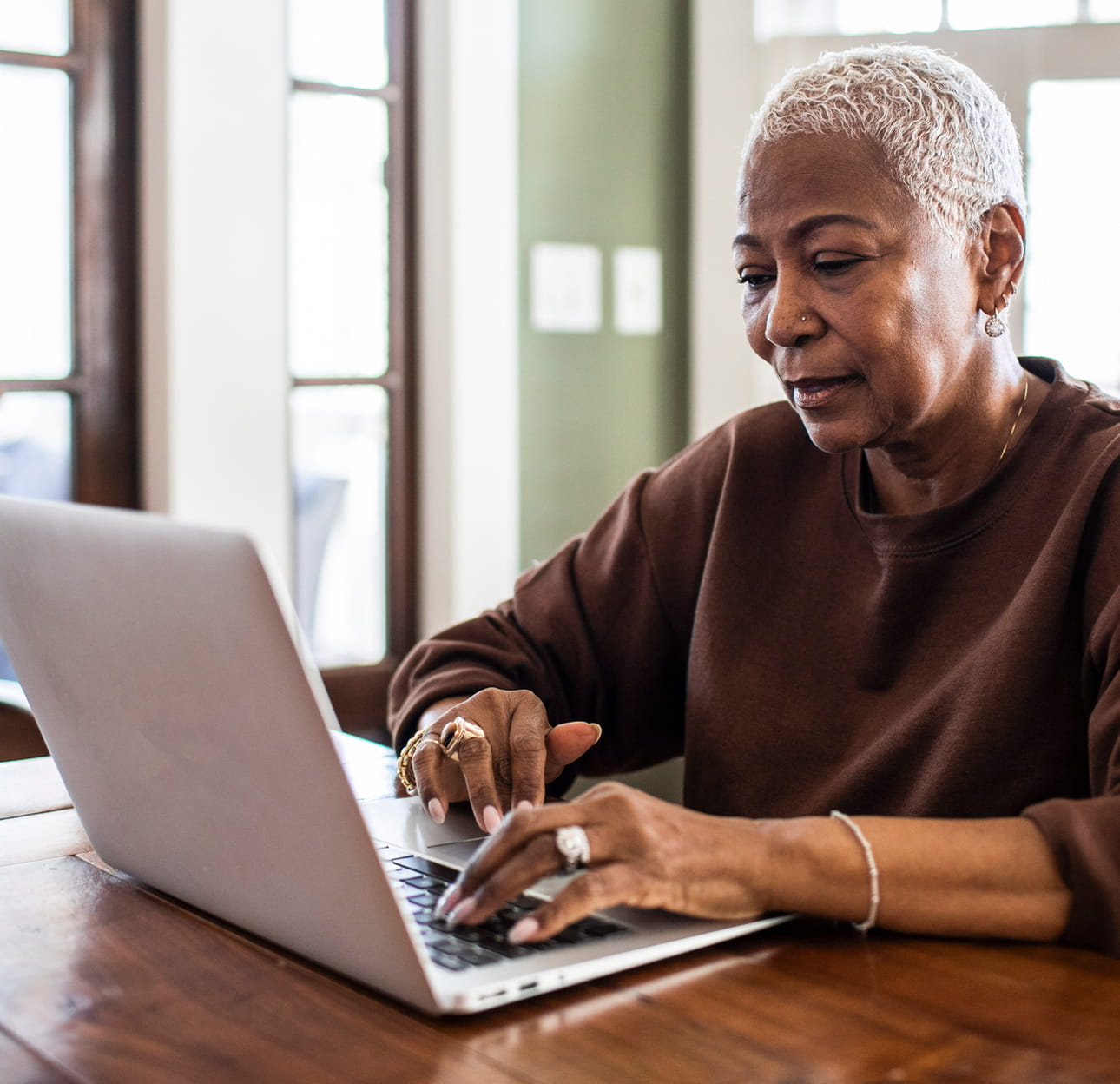 An African-American woman with short blonde hair is wearing a brown sweatshirt. She is typing on a keyboard, looking at a laptop screen with a neutral expression.