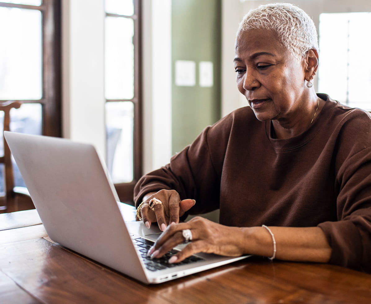 An African-American woman with short blonde hair is wearing a brown sweatshirt. She is typing on a keyboard, looking at a laptop screen with a neutral expression.