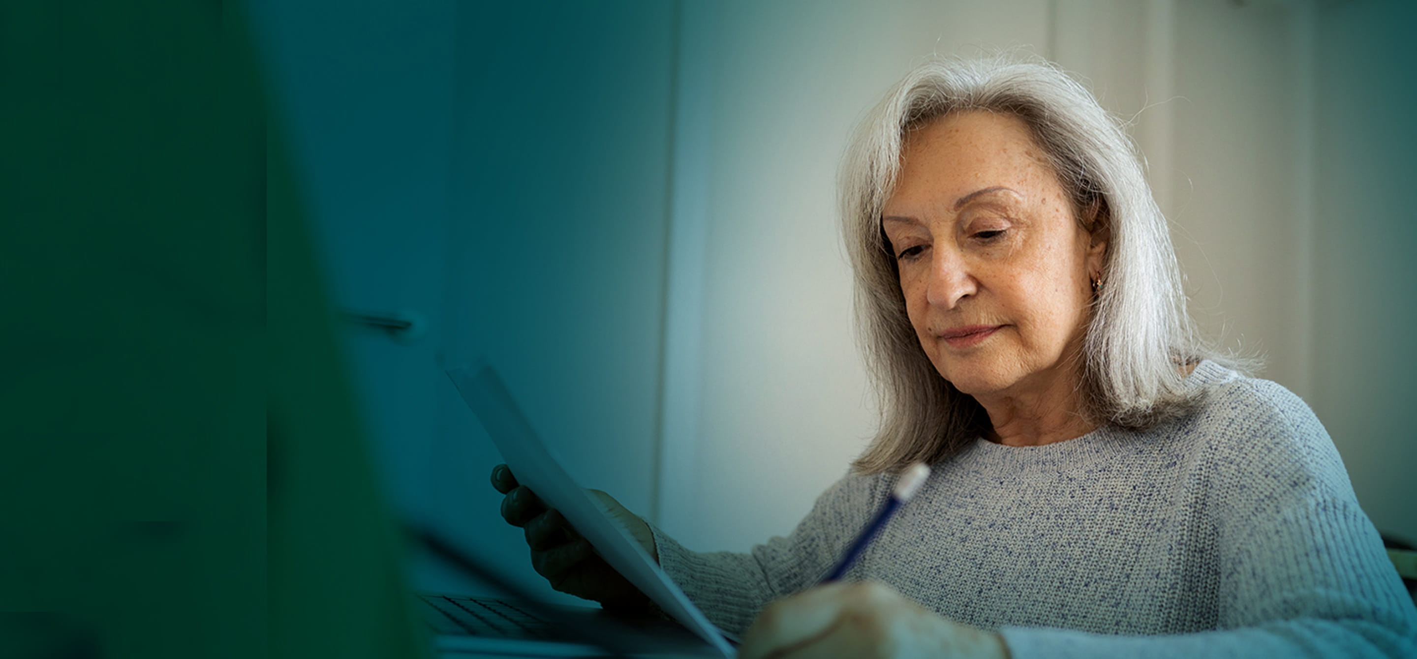 A senior woman sitting in front of a laptop, looking down at a paper and writing with a pencil.