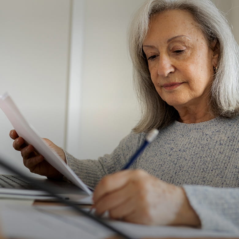 A senior woman sitting in front of a laptop, looking down at a paper and writing with a pencil.